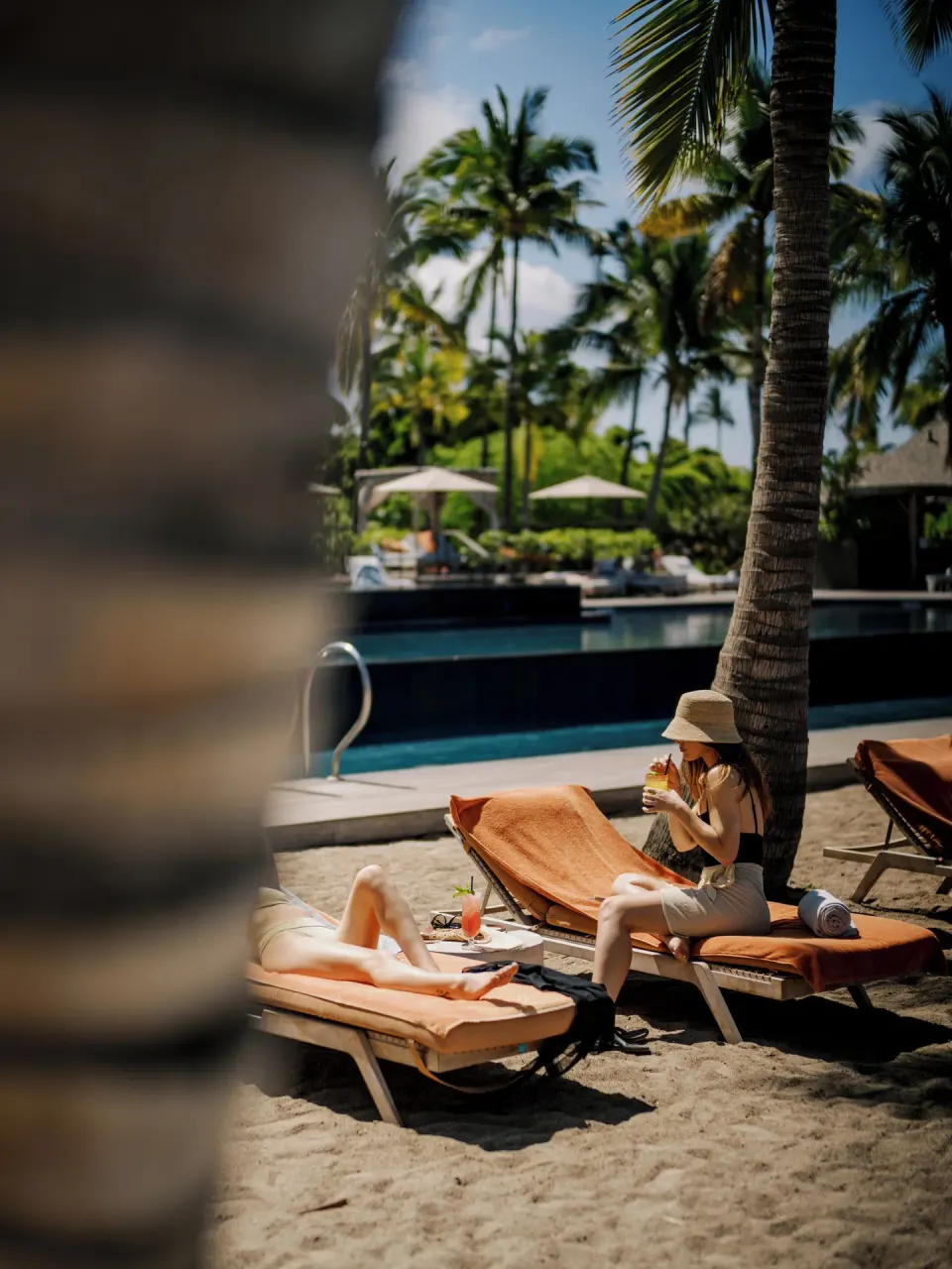 Guests relaxing on sandy poolside loungers beneath palms at Kona Village.
