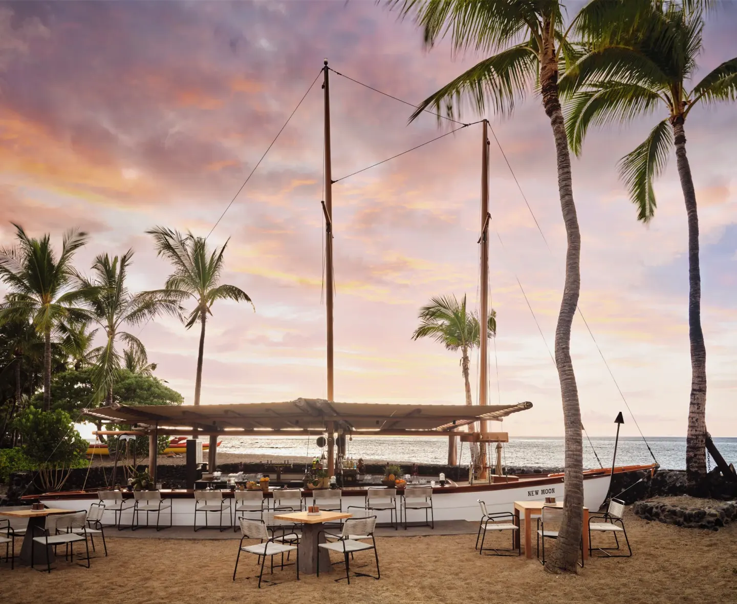 A sailboat-style beach bar with tables sits under palms at sunset at Kona Village.