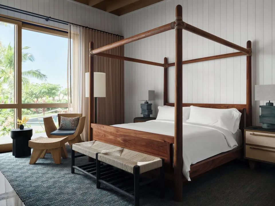 Four-poster bed beside large windows in an Ohana Pool Kauhale room at Kona Village.
