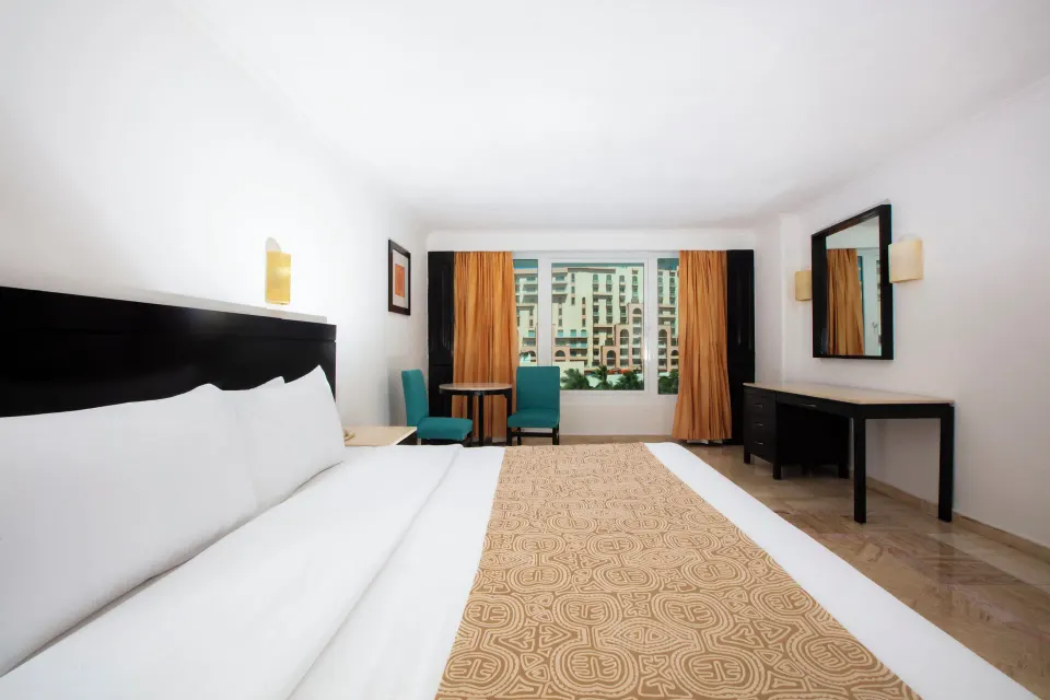 Standard Room king bed facing teal chairs, desk, mirror, and city view window at Krystal Cancun.