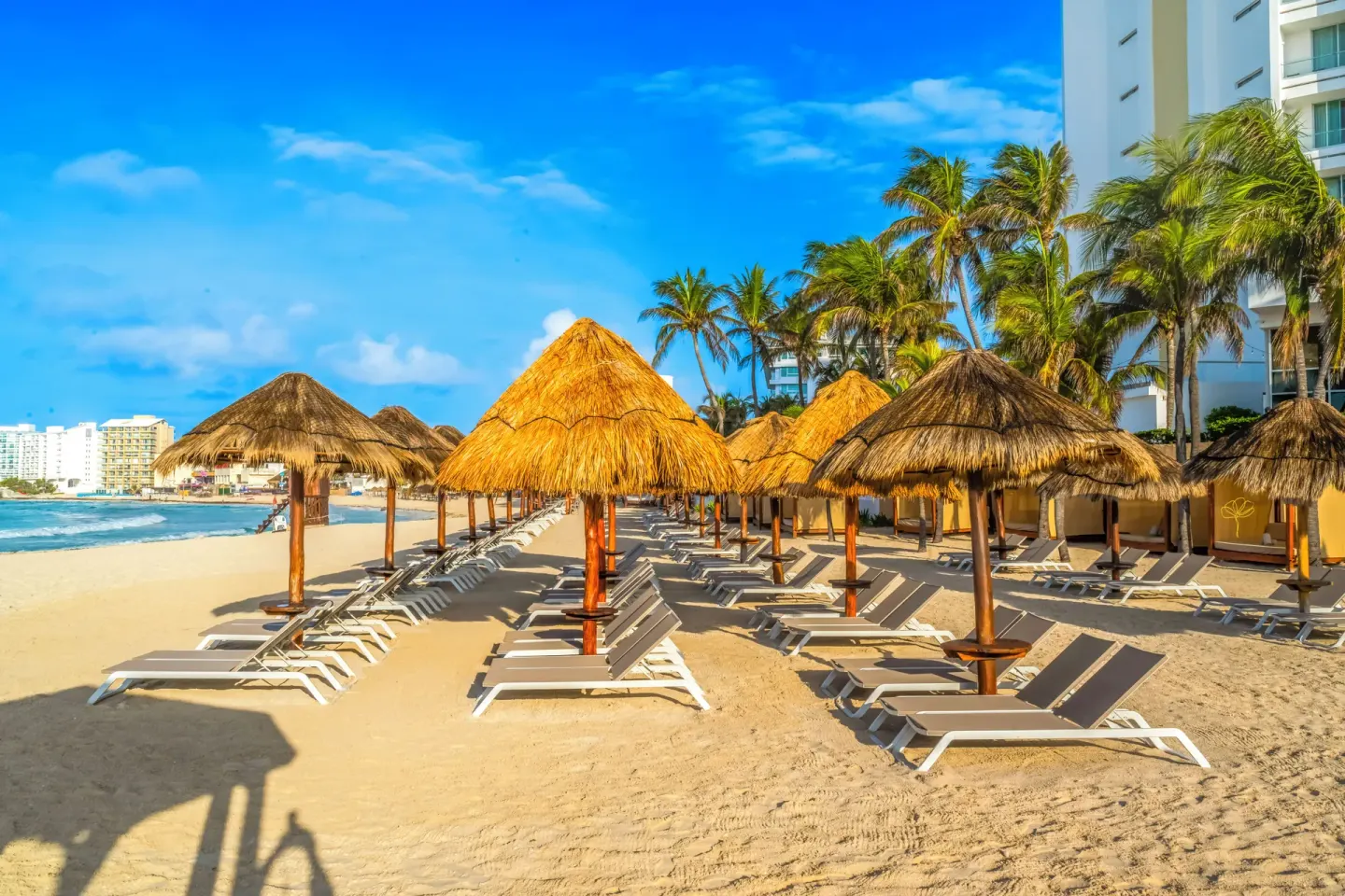 Beach loungers under thatched palapas beside the hotel tower at Krystal Grand Cancun.