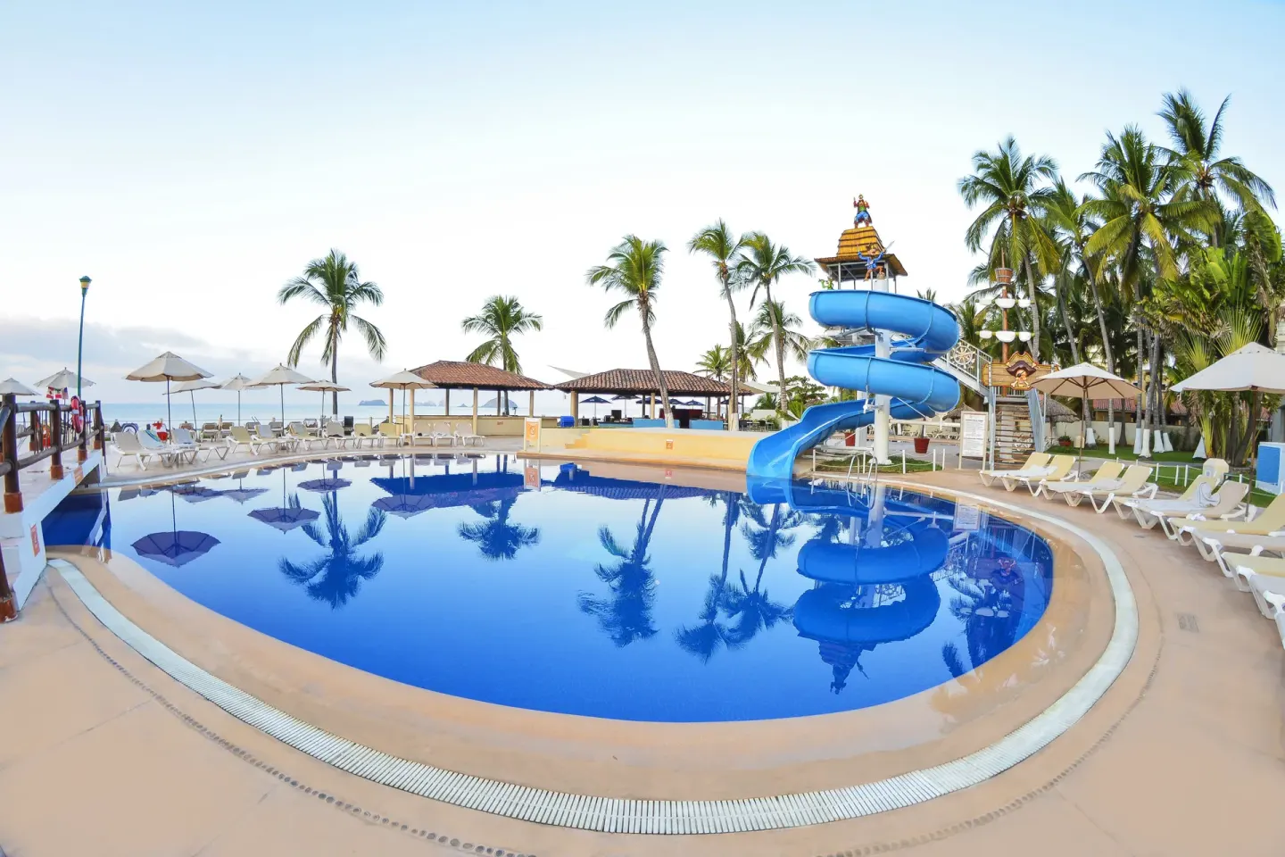 Daytime pool with a blue waterslide, palms, loungers, and ocean backdrop at Krystal Ixtapa.