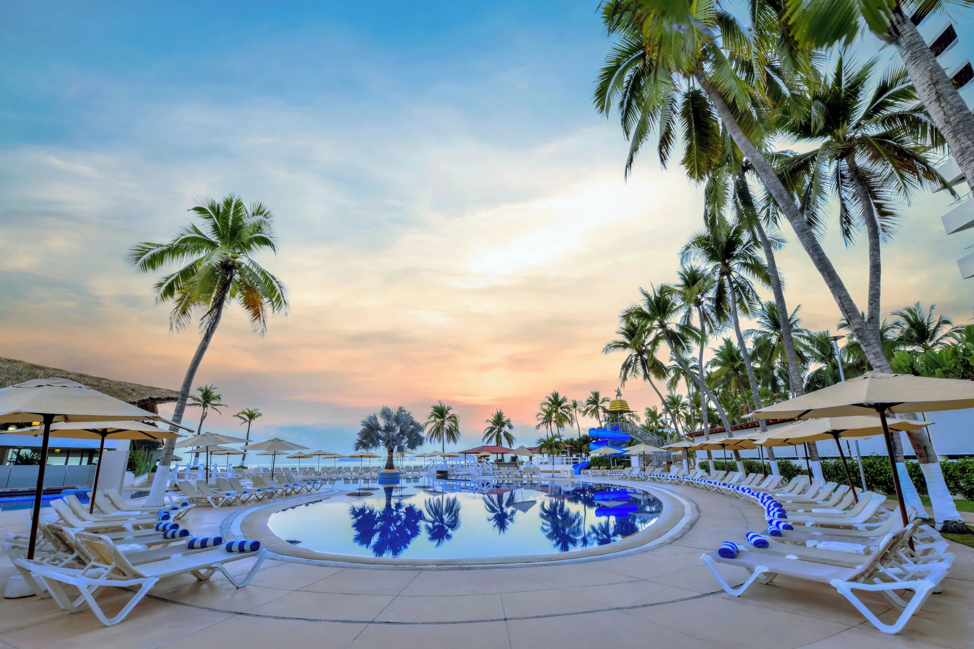 Curved pool at sunset with loungers, palms, umbrellas, and waterslide at Krystal Ixtapa.
