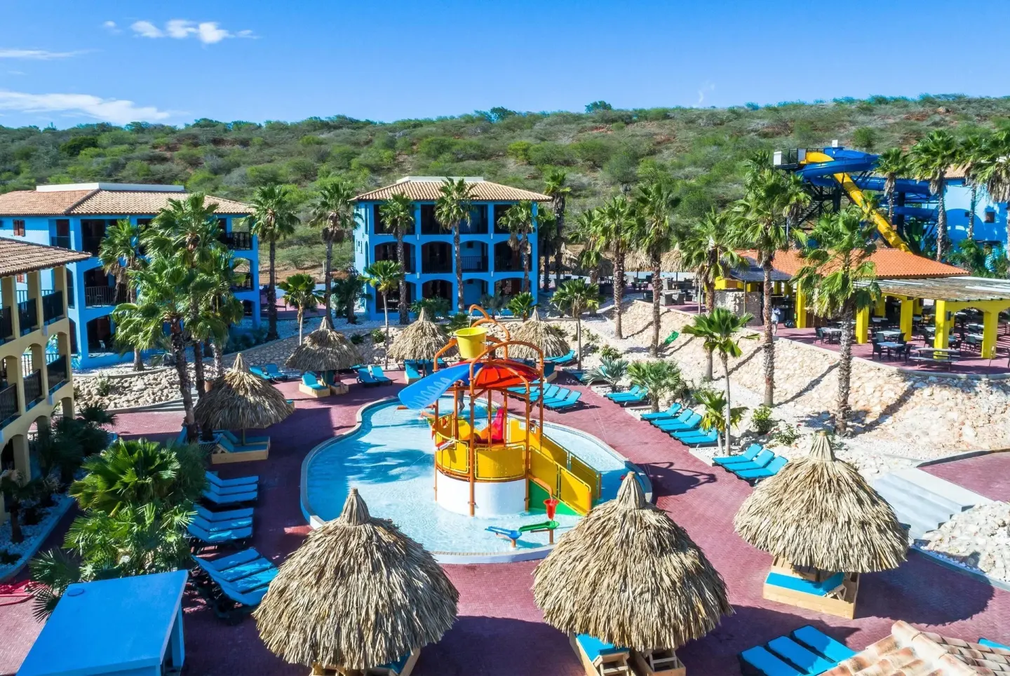 Aerial view of the pool, slides, palapas, and blue buildings at Kunuku Resort Curacao.