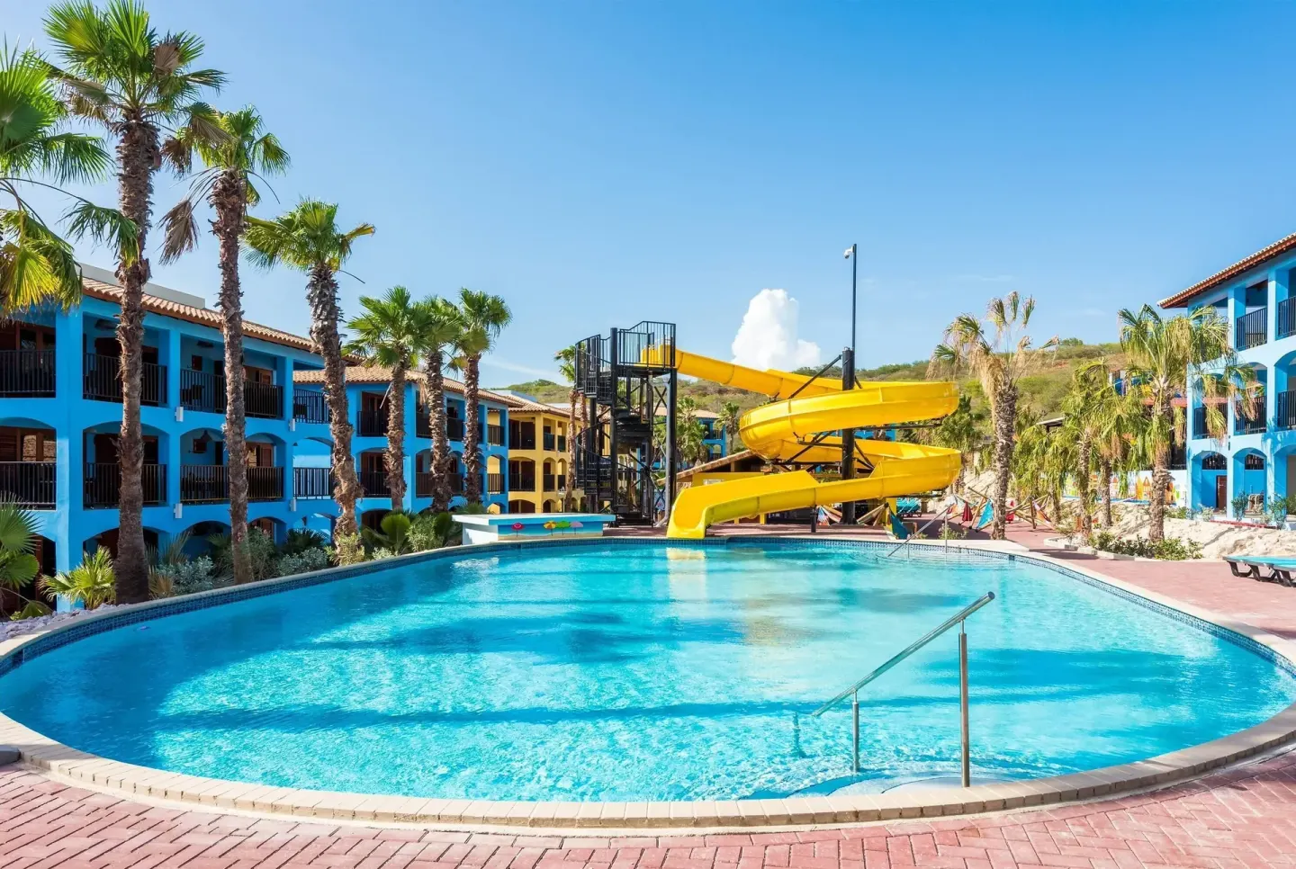 Large pool with a yellow spiral waterslide and colorful buildings at Kunuku Resort Curacao.