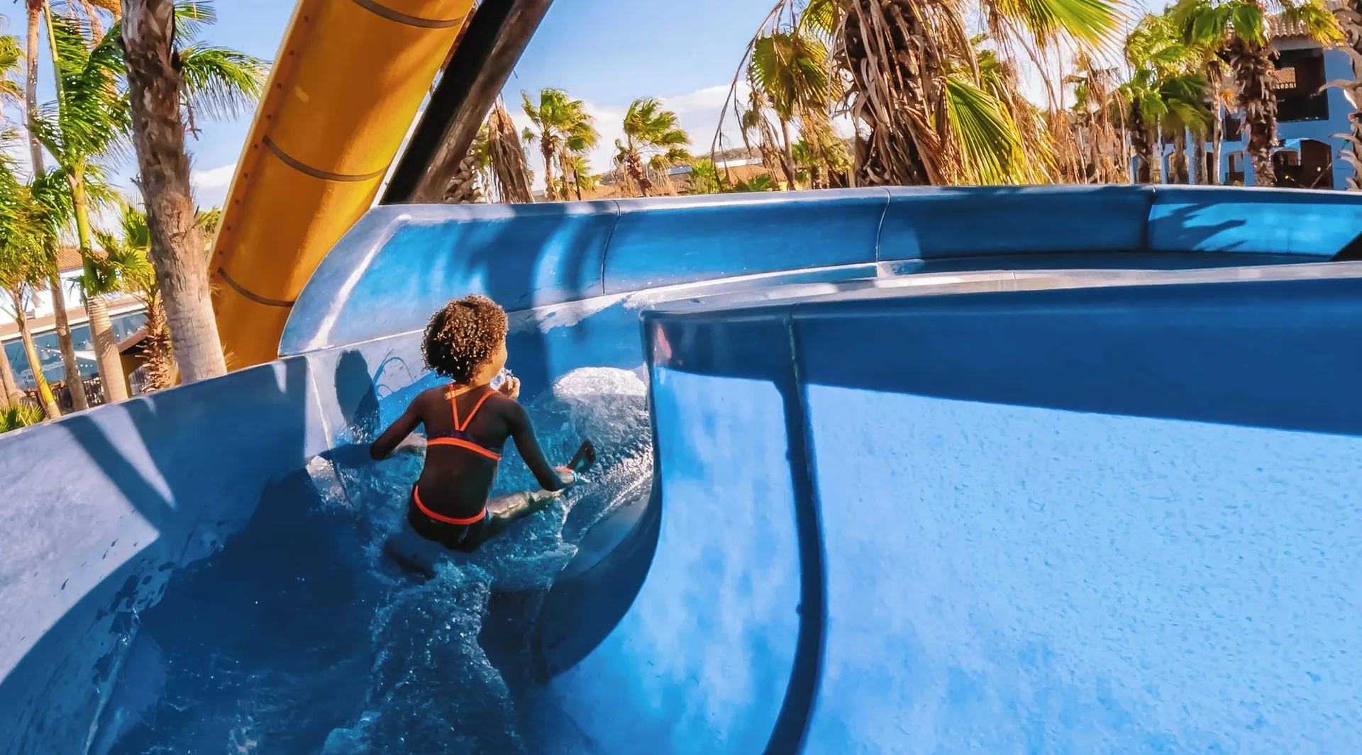 Child rides a blue waterslide curve lined with palm trees at Kunuku Resort Curacao.