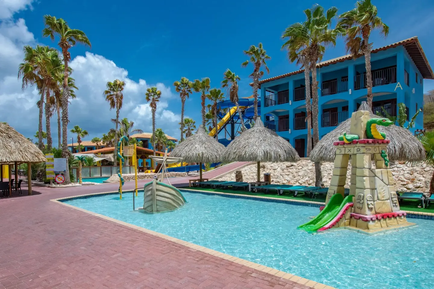 Pool area with small slides, thatched umbrellas, and blue buildings at Kunuku Resort Curacao.