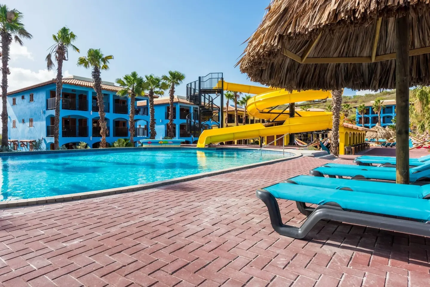 Pool loungers under a palapa beside a yellow waterslide at Kunuku Resort Curacao.