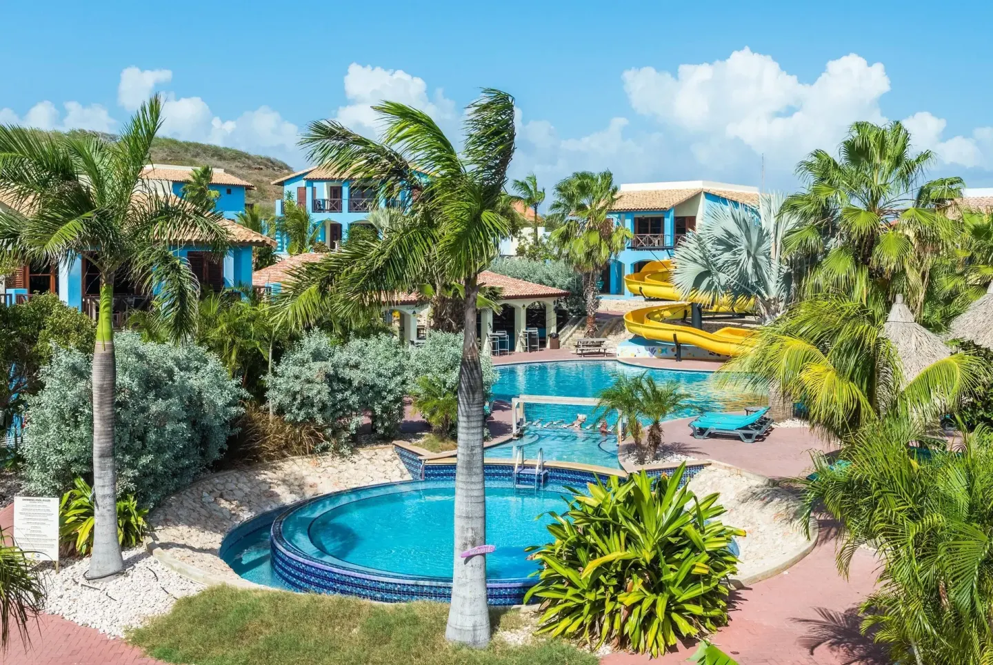 Tropical resort pools, palms, blue buildings, and a yellow slide at Kunuku Resort Curacao.