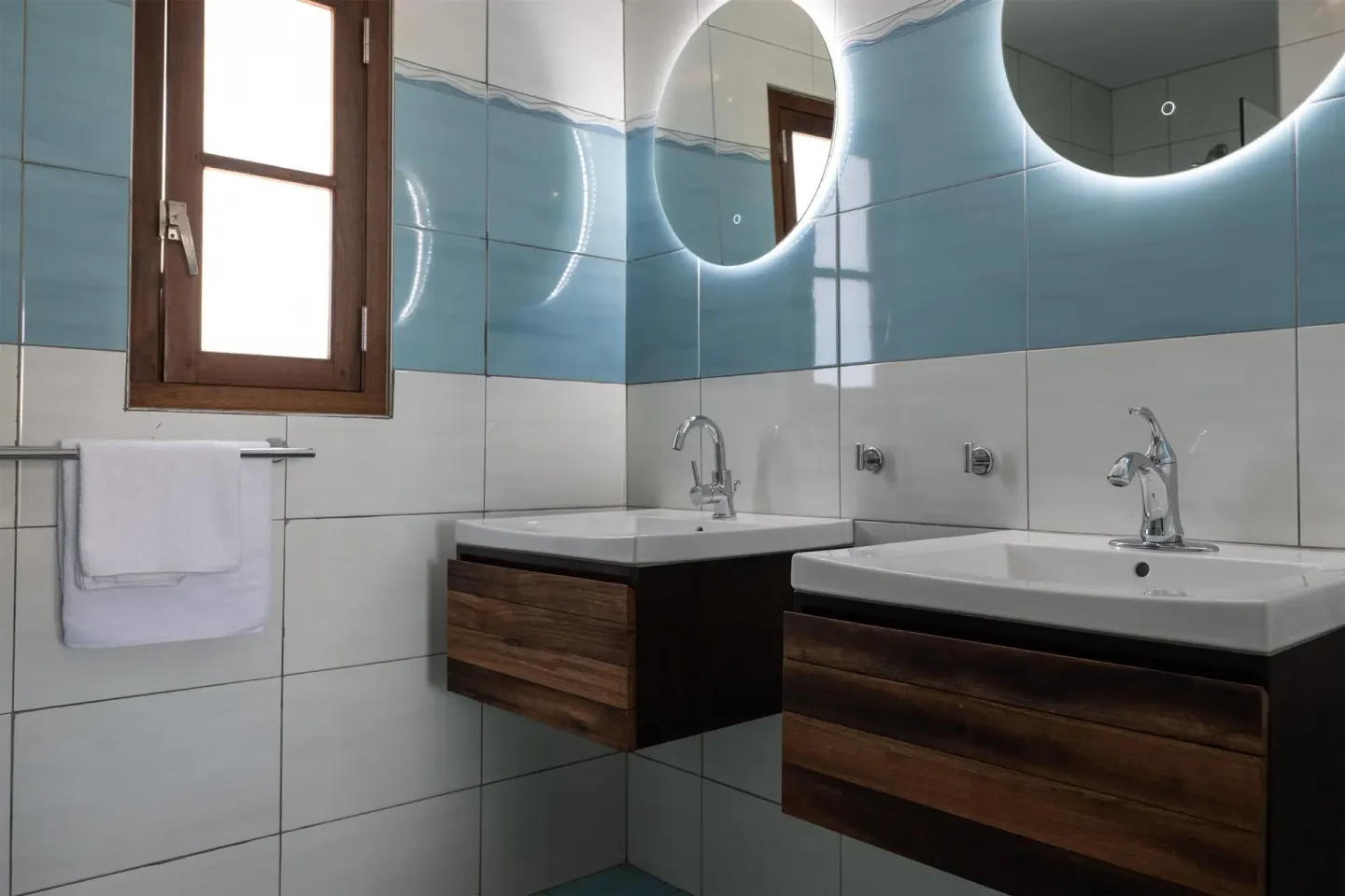 Bathroom with twin sinks, round lit mirrors, and blue tile at Kunuku Resort Curacao.