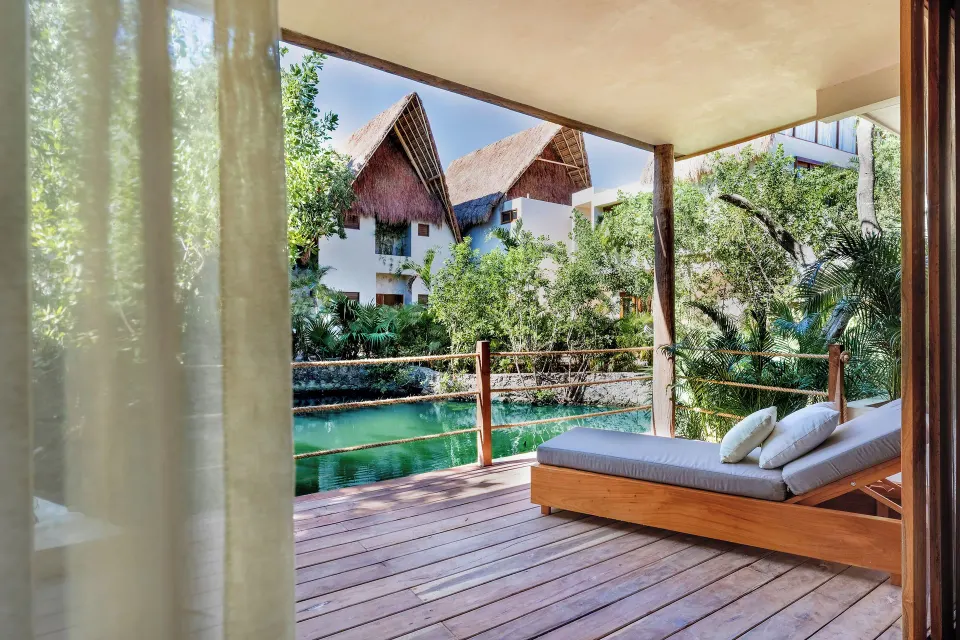 Wooden deck with a lounger overlooking a green lagoon and thatched buildings at La Valise Tulum.
