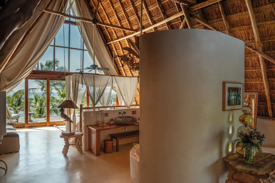 Interior view of a palapa suite showing a canopy bed, curved privacy wall and ocean-facing windows at La Valise Tulum.