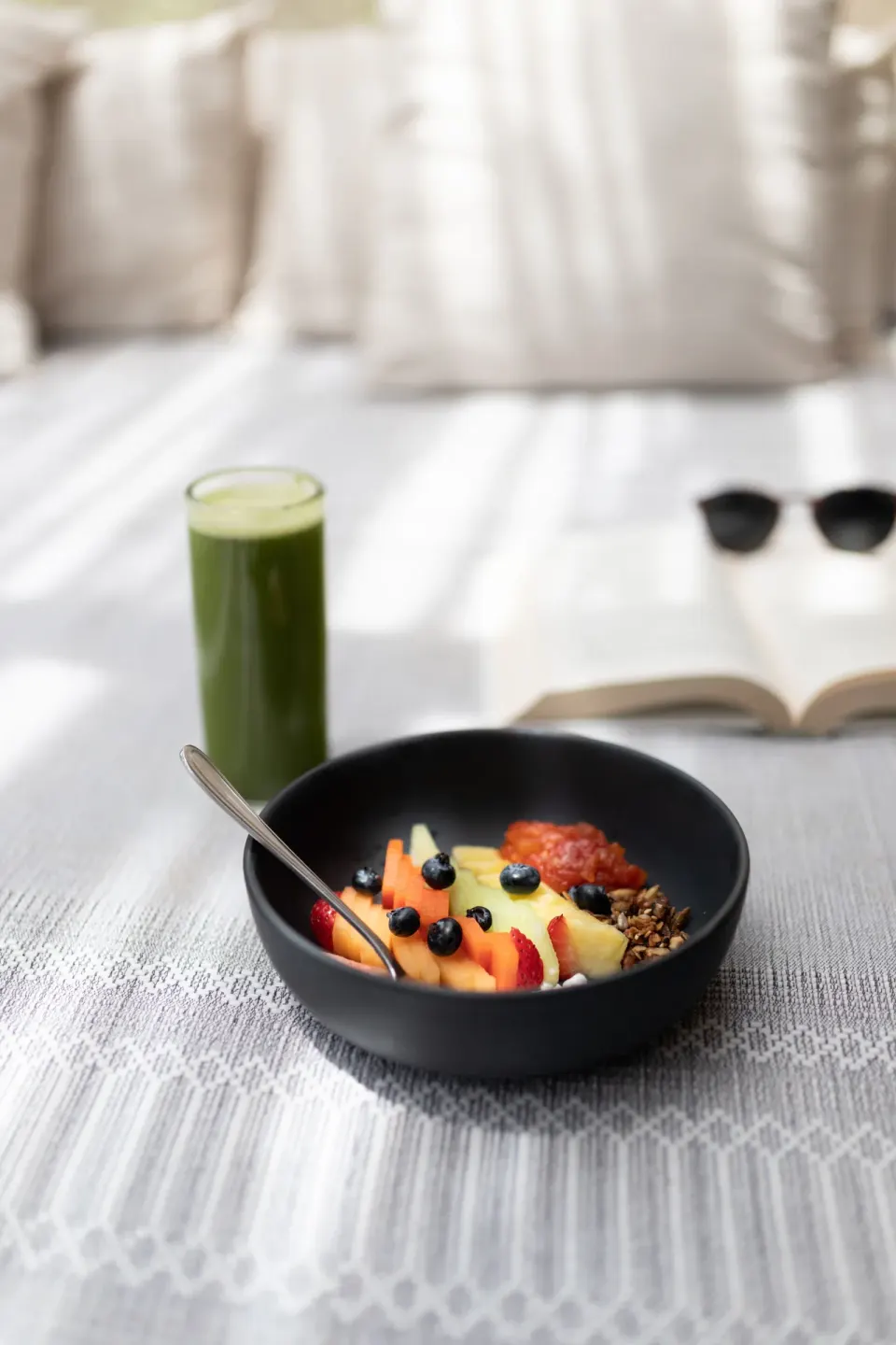 Bowl of fruit and granola with green juice and book on a bed at La Valise Tulum