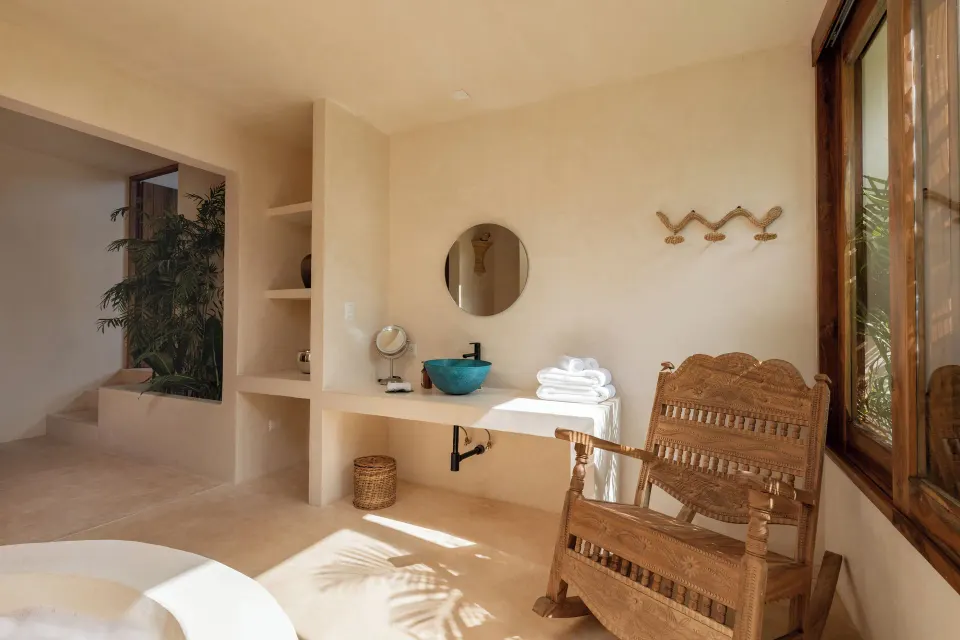 Bathroom vanity area with woven chair and round mirror in a sunlit room at La Valise Tulum