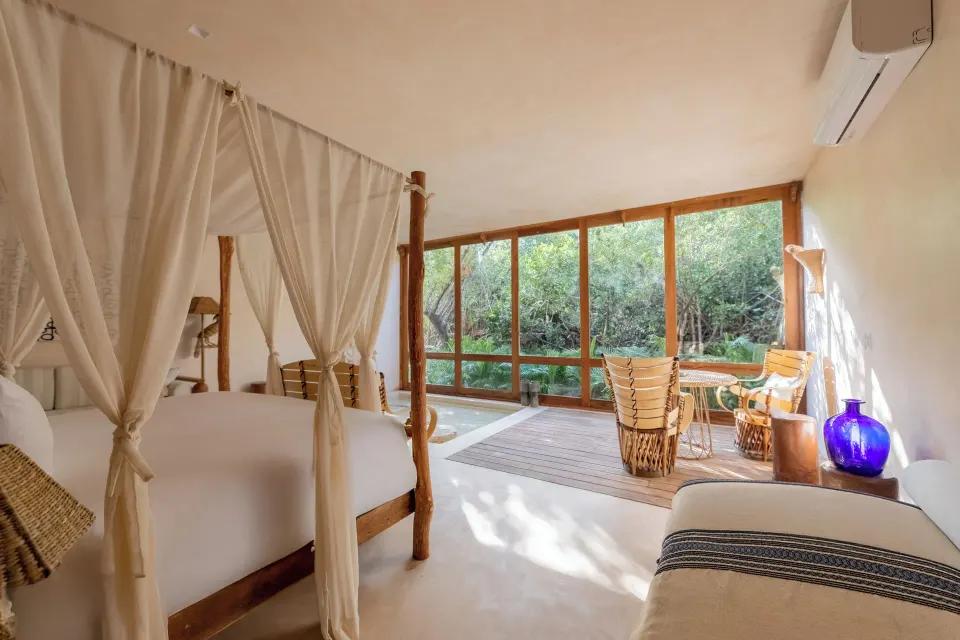 Four-poster bed and seating area facing glass wall with jungle view in a Signature Suite at La Valise Tulum