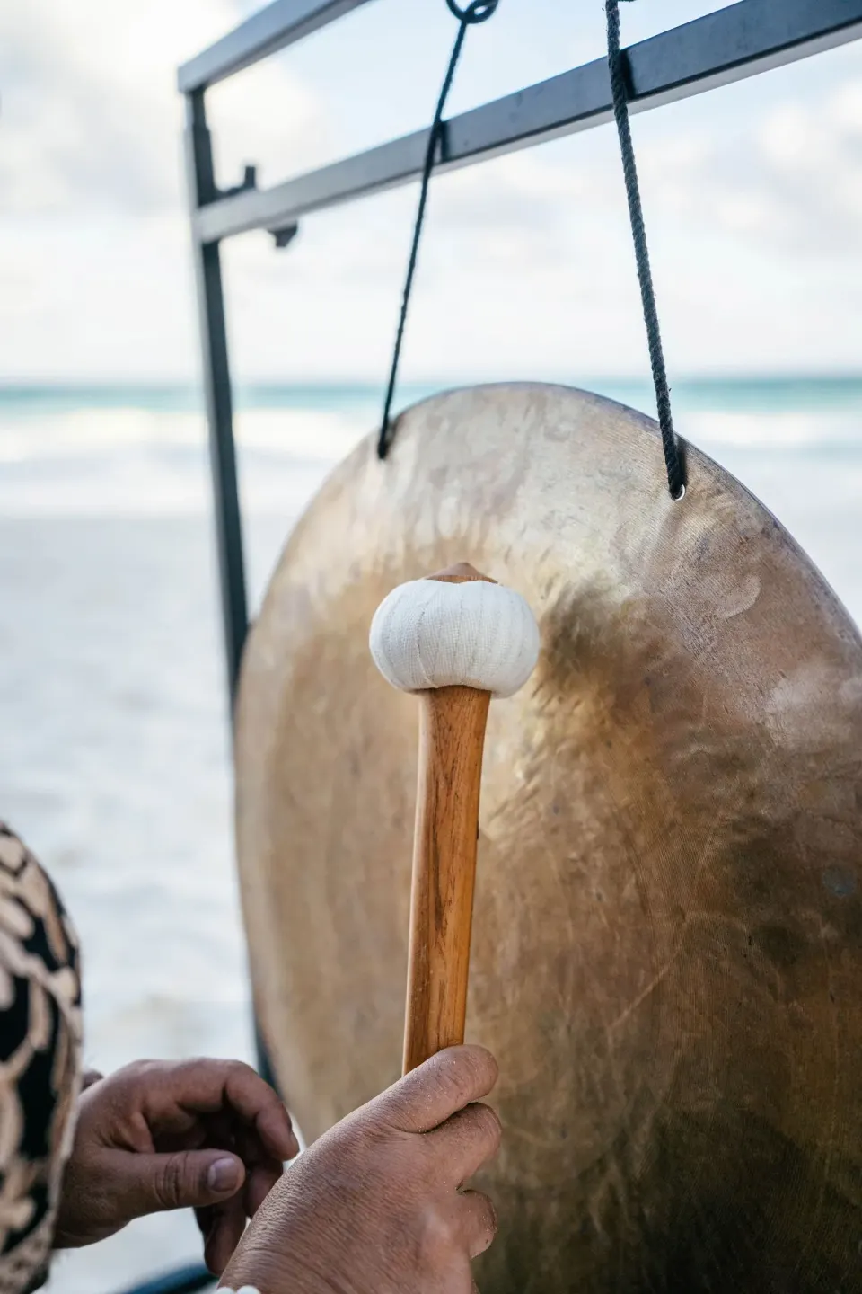 Hand holding a mallet striking a hanging bronze gong on the beach at La Valise Tulum.