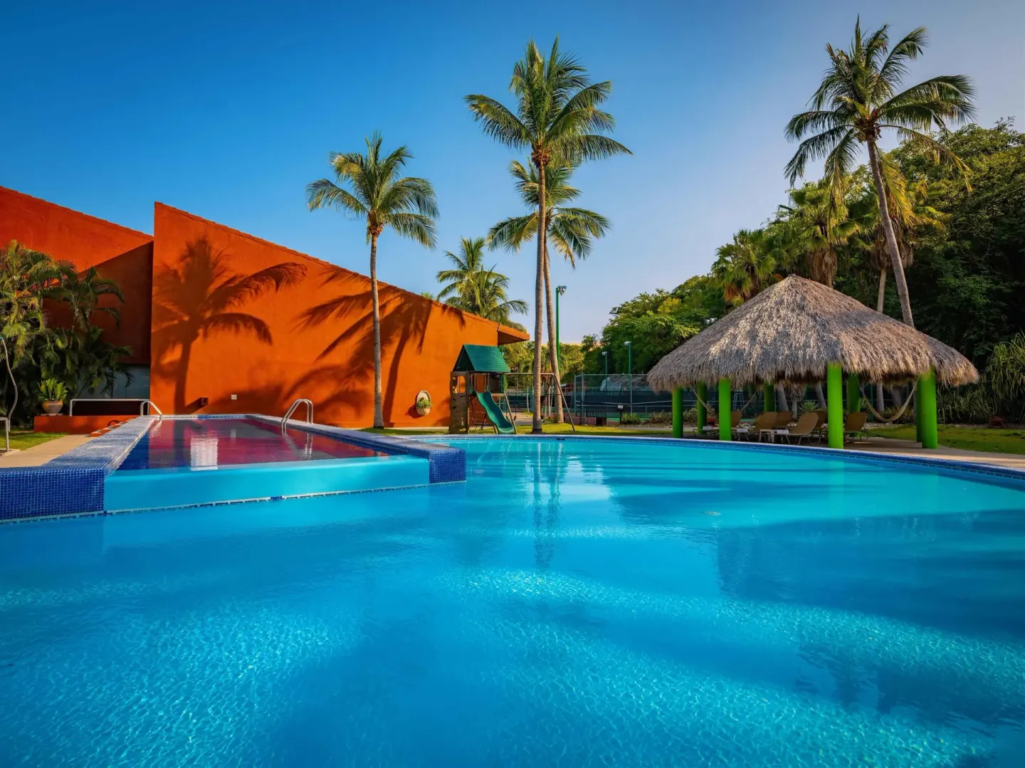 Resort pool with orange walls, palms and a thatched shade palapa at Las Brisas Huatulco.