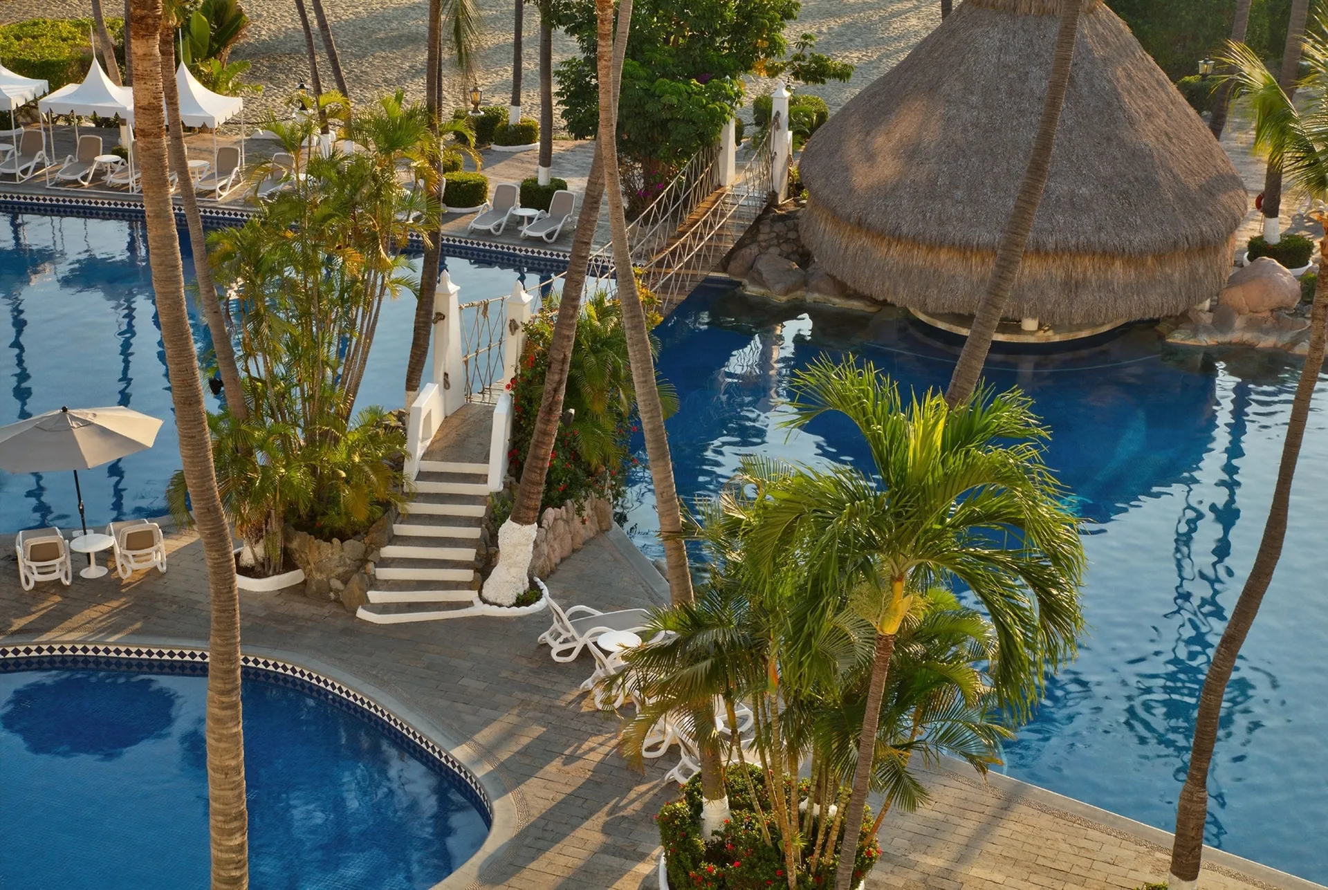 An elevated view shows connected pools, palms, loungers, and a thatched pavilion at Las Hadas.