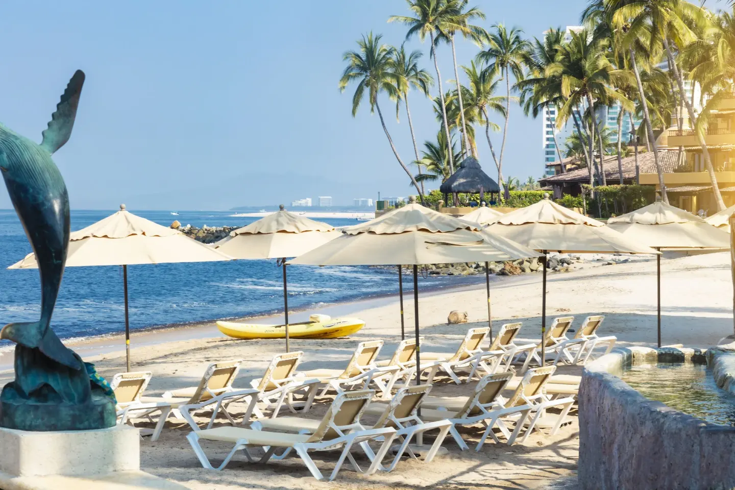 Beach seating at Las Palmas by the Sea with umbrellas, palms, a dolphin sculpture, and ocean beyond.
