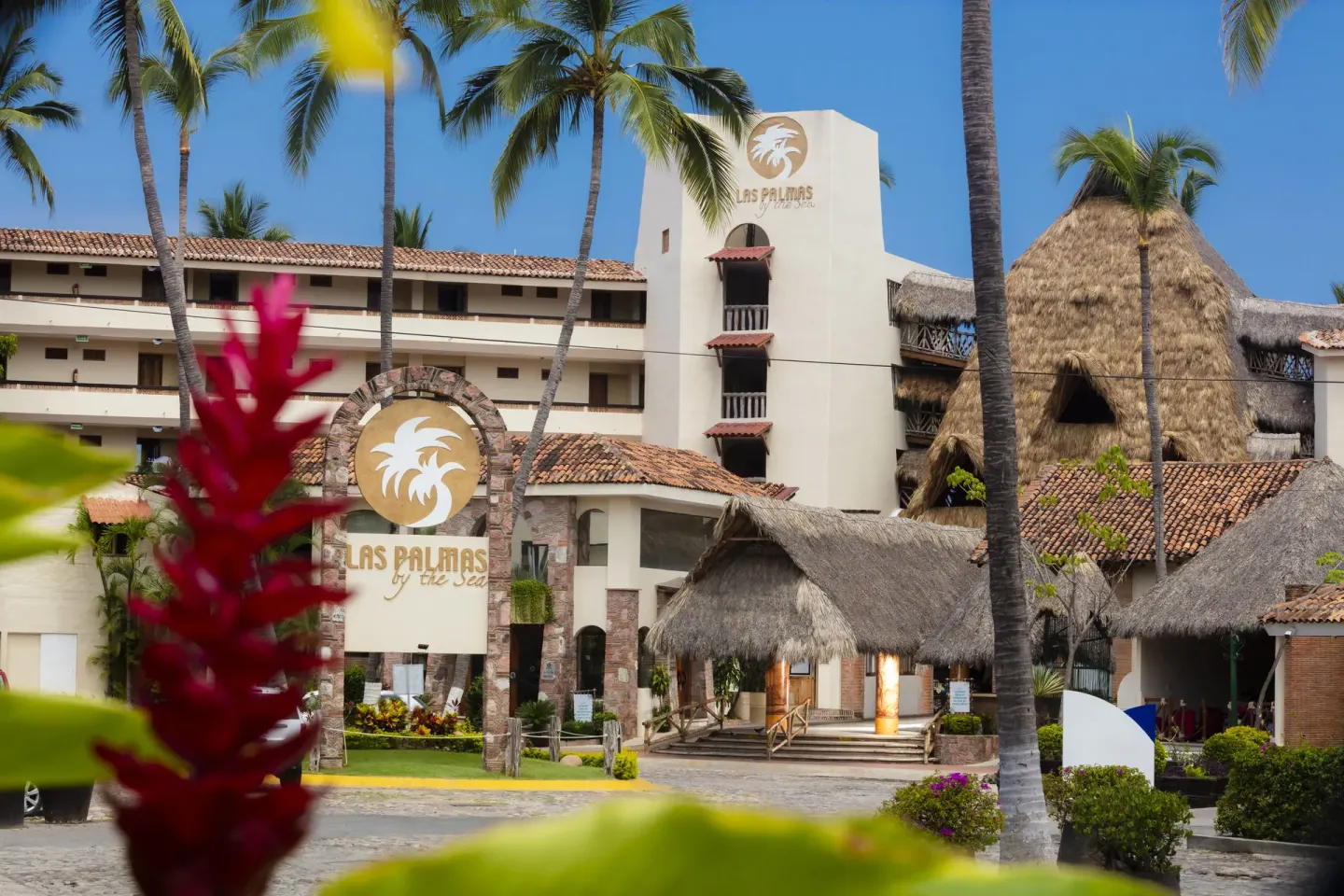 Daytime exterior with entrance sign, palms, and thatched roofs at Las Palmas by the Sea.