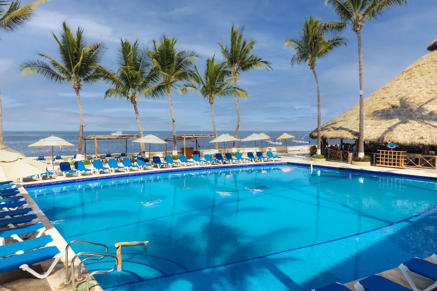 Large pool at Las Palmas by the Sea lined with blue loungers, palms, umbrellas, and ocean views.