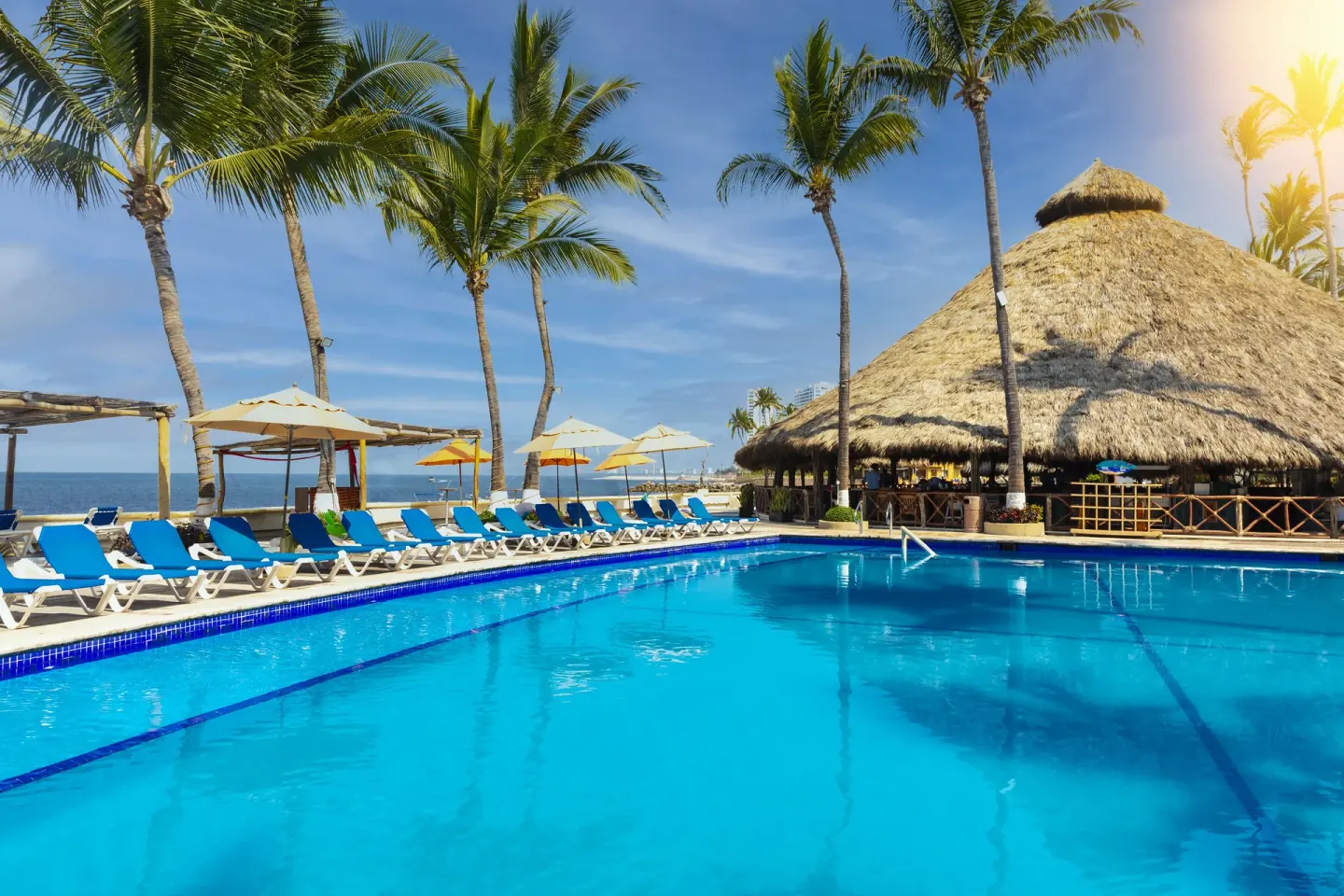Pool at Las Palmas by the Sea with blue loungers, palm trees, ocean views, and a thatched pavilion.