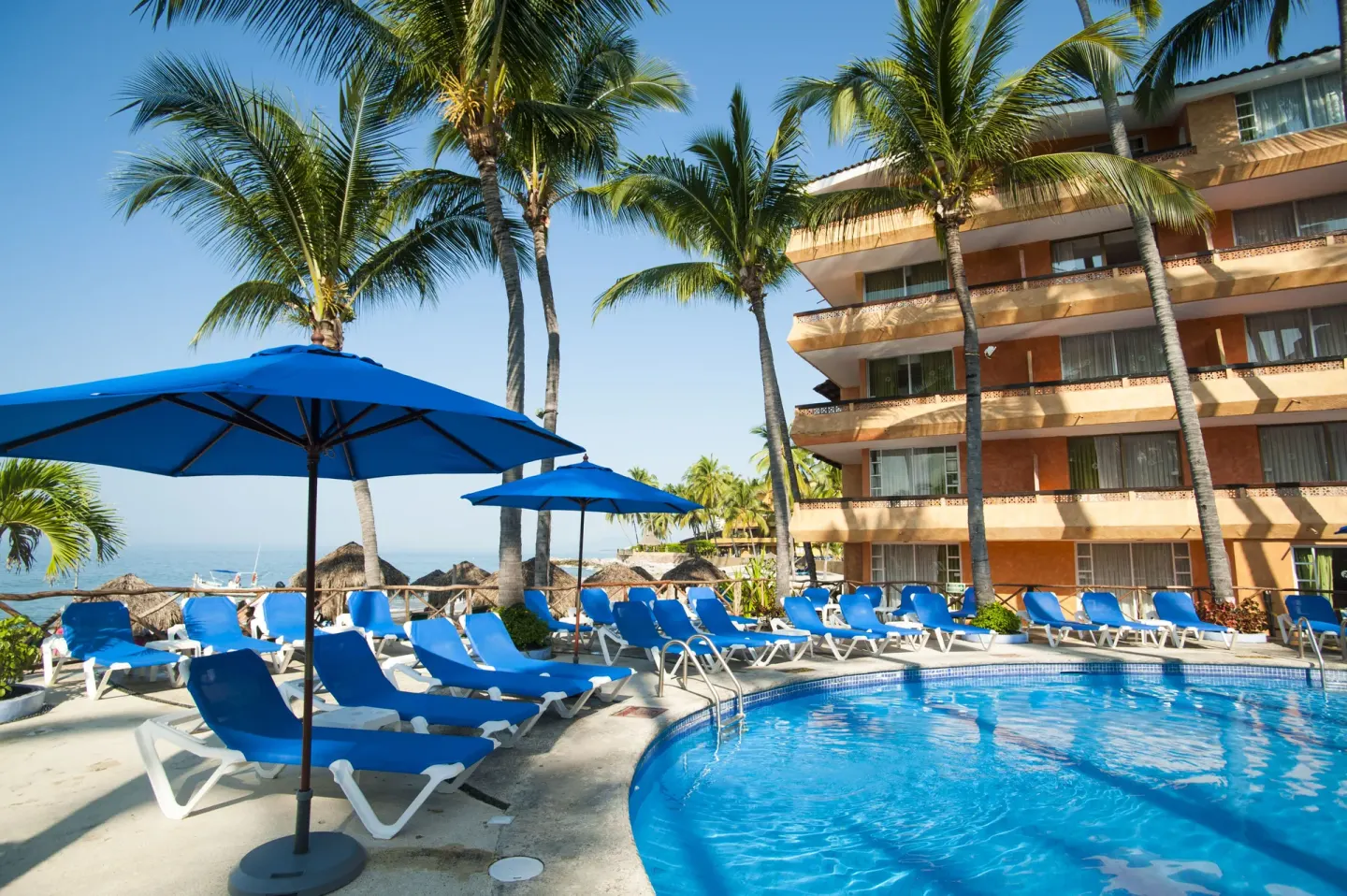 Pool deck at Las Palmas by the Sea with blue umbrellas, loungers, palms, and guestroom balconies.