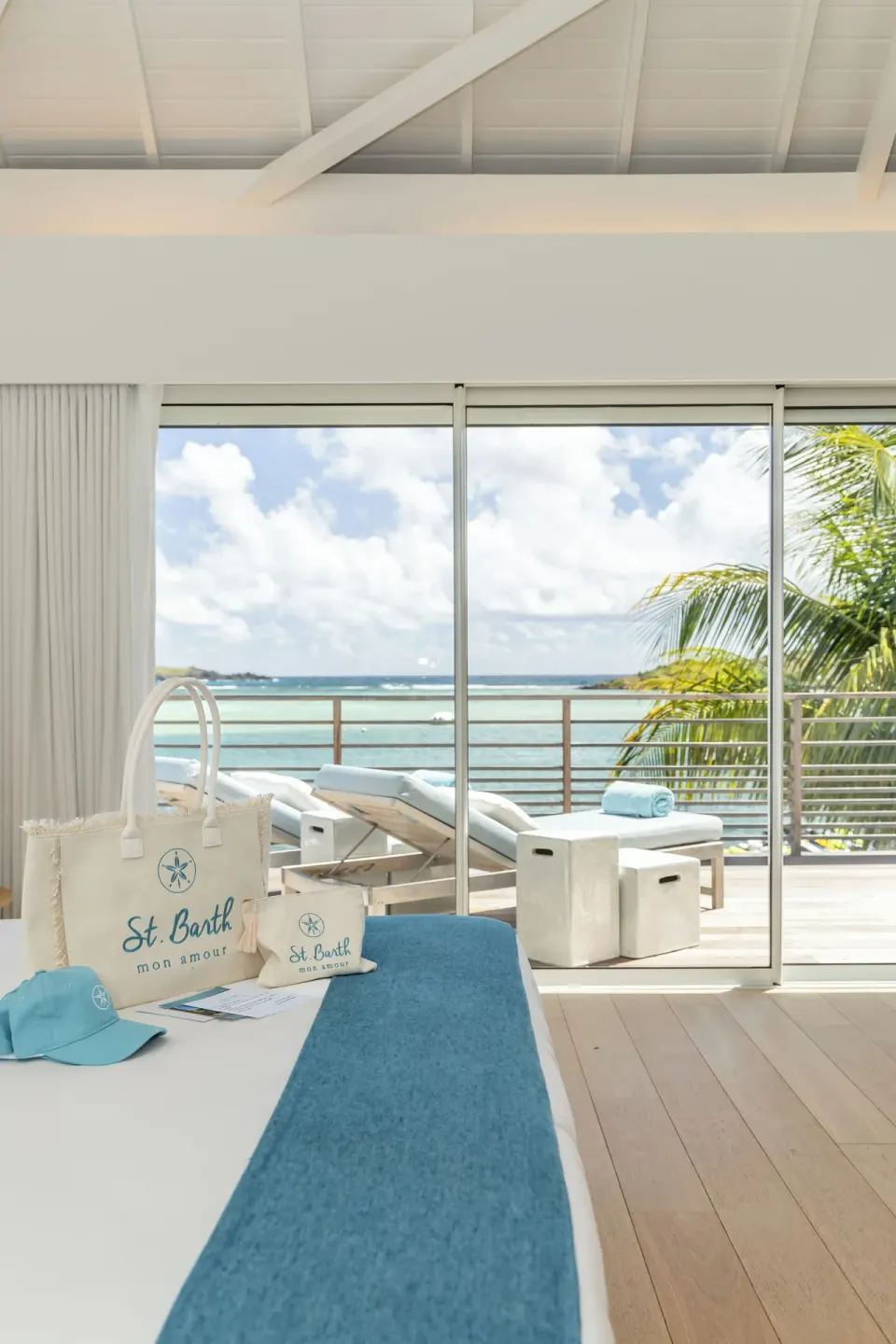 Guest room bed with St. Barth items and balcony loungers overlooking the sea at Le Barthélemy Hotel & Spa.