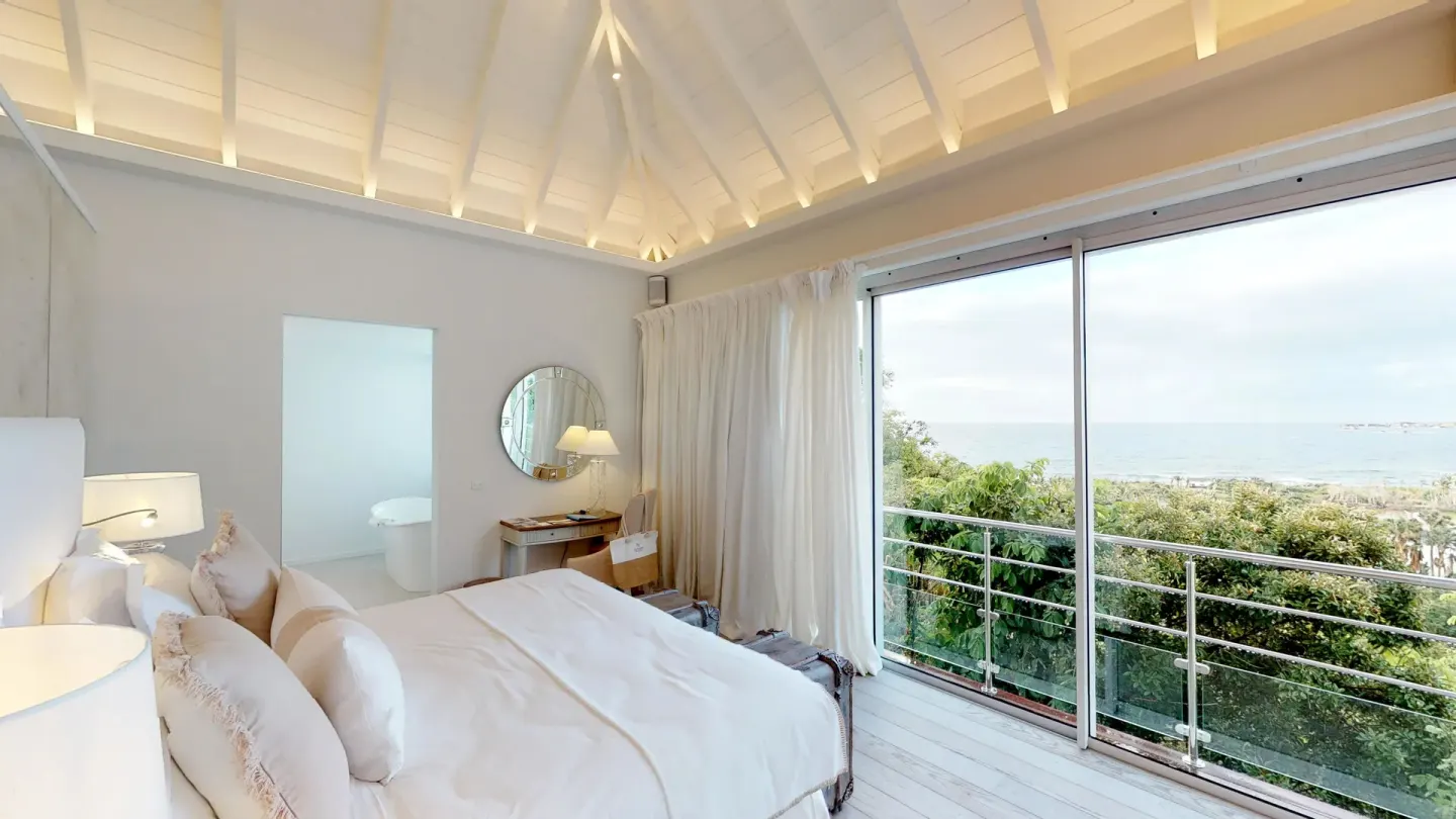 Guest room with a white bed, vaulted ceiling, balcony, and ocean view at Le Toiny Hôtel & Beach Club.
