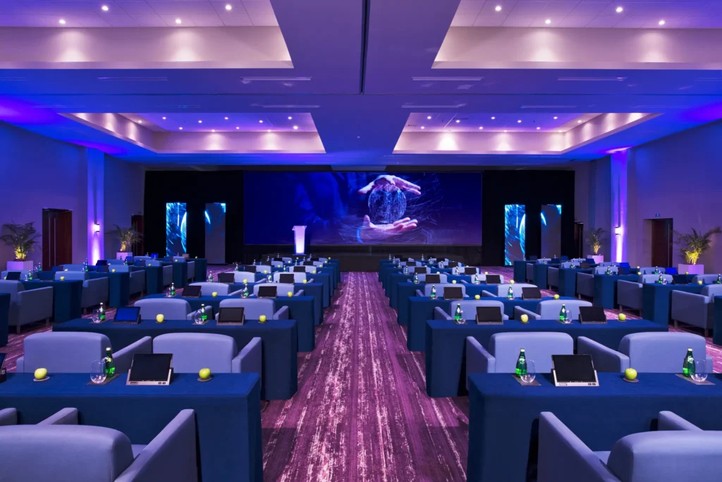 Ballroom arranged in a classroom-style layout with blue draped tables, tablets, bottled water, and stage at Breathless Riviera Cancun.