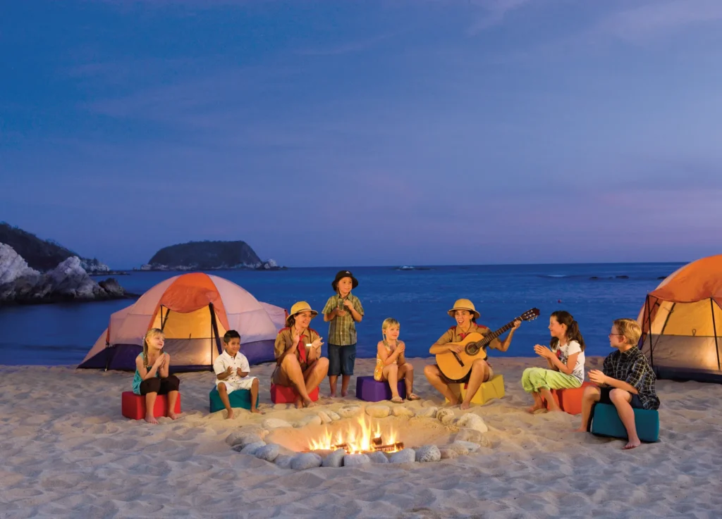 Family beach camp-out experience with bonfire at Dreams Huatulco Resort & Spa under the evening sky.