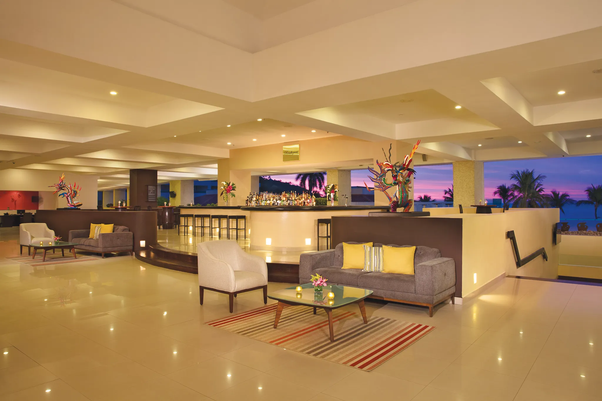 Rendezvous Lobby Bar at Dreams Huatulco Resort & Spa featuring elegant lounge seating, a stylish central bar, and sunset ocean views.