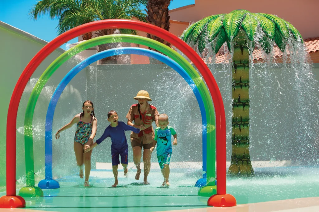Kids running through colorful water spray arches at the splash park at Dreams Los Cabos Suites Golf Resort & Spa, a fun family water play area in Los Cabos.