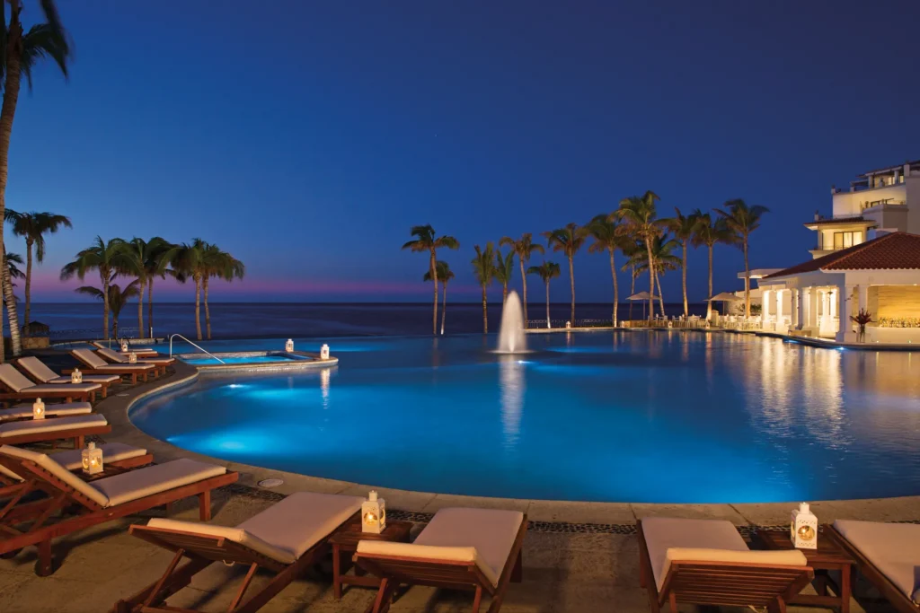 Illuminated resort pool and lounge chairs at night overlooking the ocean at Dreams Los Cabos Suites Golf Resort & Spa.