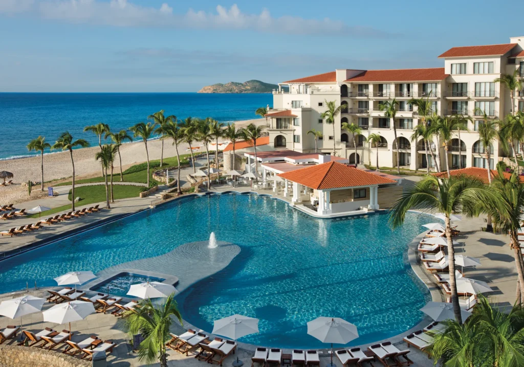 Panoramic view of the oceanfront main pool surrounded by palm trees at Dreams Los Cabos Suites Golf Resort & Spa.