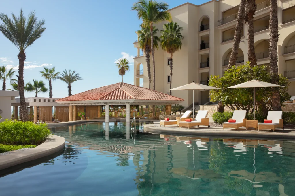 Manatees swim-up pool bar at Dreams Los Cabos Suites Golf Resort & Spa featuring a shaded palapa bar in a tropical resort pool with lounge chairs and palm trees.