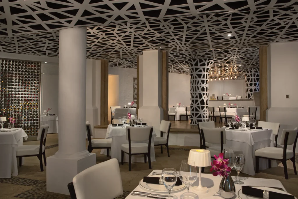 Portofino Mediterranean restaurant at Dreams Los Cabos Suites Golf Resort & Spa featuring elegant white table settings, modern lattice ceiling design, and an extensive wine display.