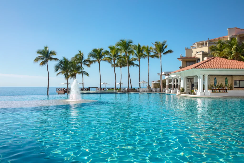 Sugar Reef pool bar at Dreams Los Cabos Suites Golf Resort & Spa featuring a tropical swim-up bar beside a large infinity-style pool with palm trees and ocean views.