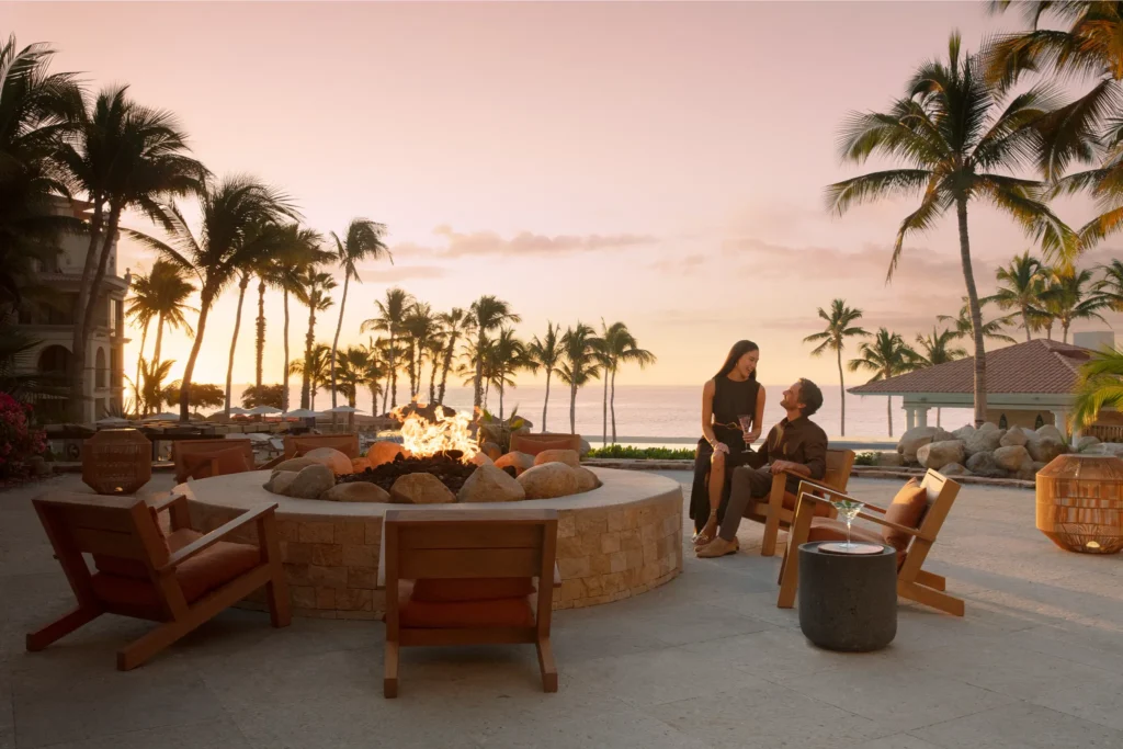 Couple enjoying sunset drinks beside an oceanfront fire pit at Dreams Los Cabos Suites Golf Resort & Spa.