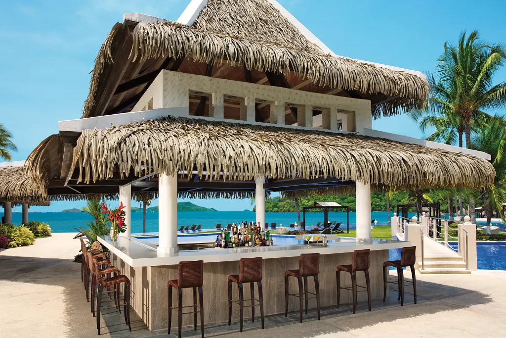 Barracuda Pool Bar at Dreams Playa Bonita Panama featuring a thatched-roof bar, poolside seating, and ocean views near the beach.