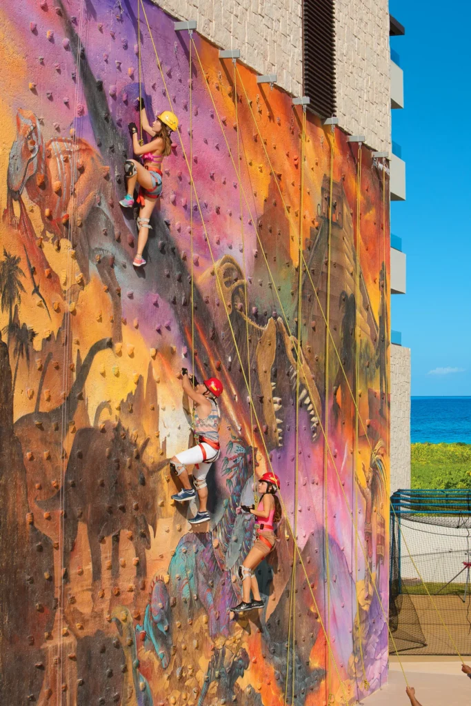 Kids climbing a colorful outdoor rock wall at Dreams Playa Mujeres Resort with ocean views.