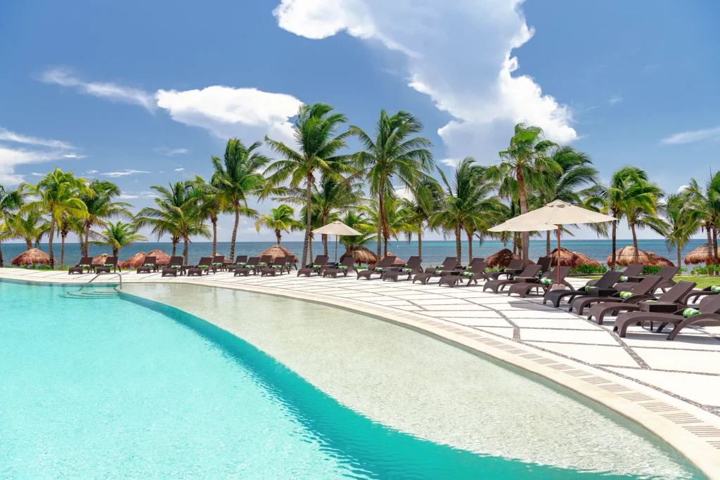 Curved infinity-style pool lined with loungers, umbrellas, and palm trees by the beach.