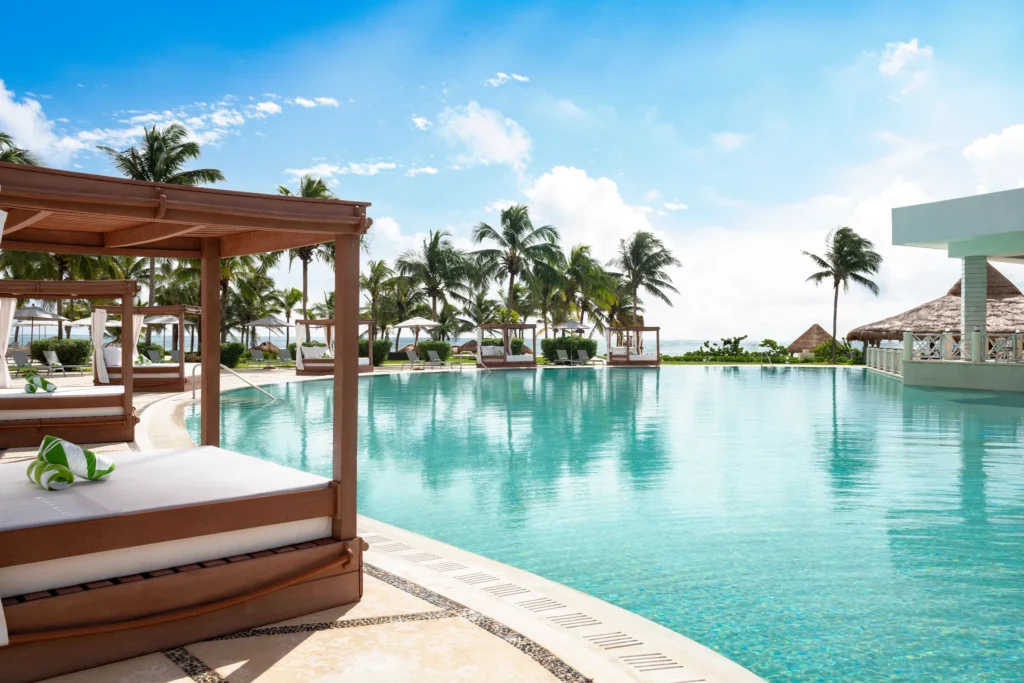 Large resort pool with cabanas and palm trees under a bright blue sky at Dreams Puerto Morelos Resort & Spa.