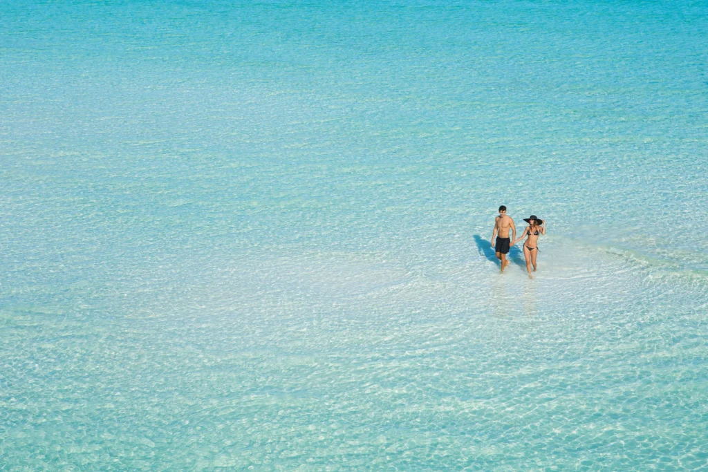 Couple walking hand in hand across shallow turquoise waters near Dreams Sands Cancun Resort & Spa.