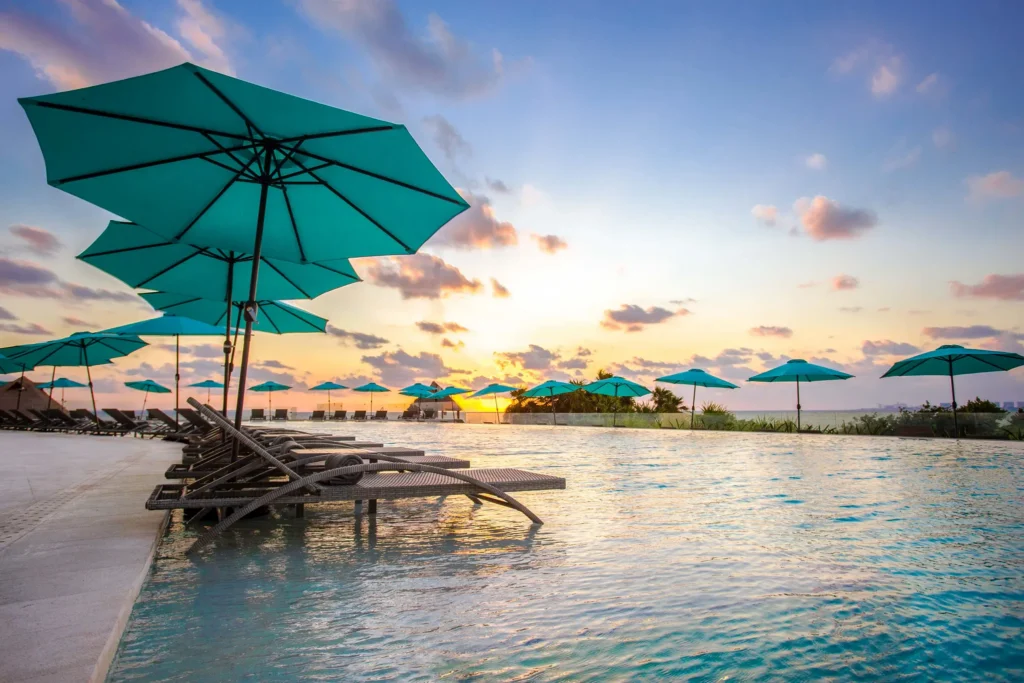 Infinity pool with lounge chairs and turquoise umbrellas at sunset at Dreams Vista Cancun Golf & Spa Resort.