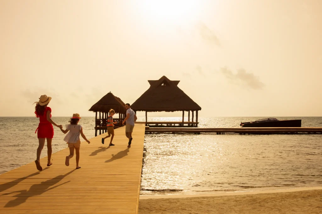 Family walking on a dock toward overwater palapas at sunset near Dreams Vista Cancun Golf & Spa Resort.