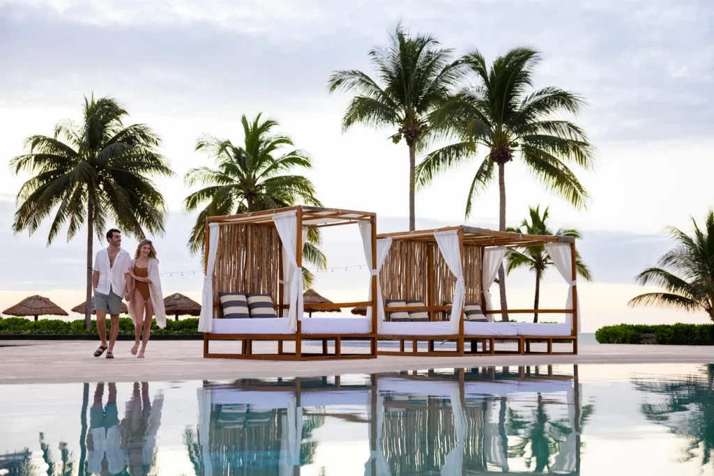 Couple walking near luxury poolside cabanas at Hyatt Zilara Riviera Maya, surrounded by palm trees and reflecting pool.