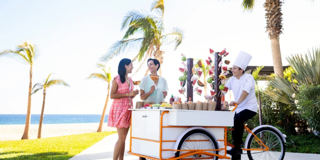 Guests enjoying fresh tropical fruit from a beachside fruit cart at Hyatt Ziva Los Cabos.