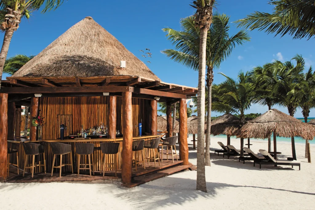 Barracuda Pool & Beach Bar at Secrets Akumal Riviera Maya featuring a palapa-style open-air bar, tropical cocktails, and beachfront lounge seating under palm trees.