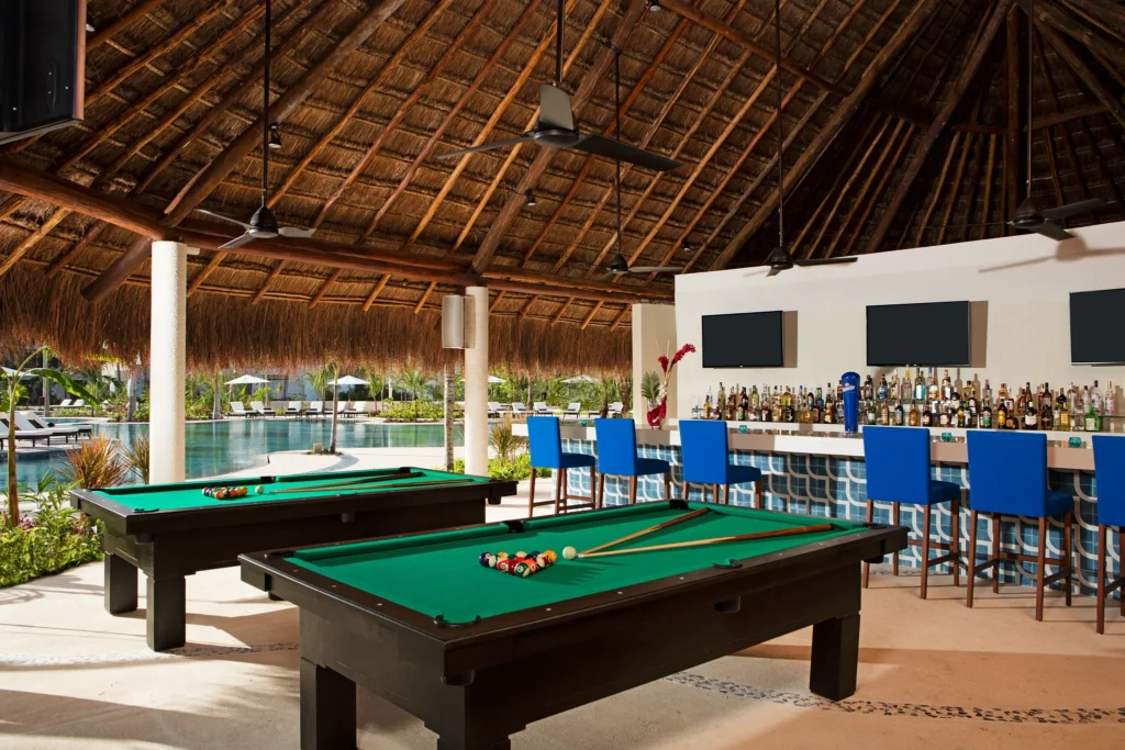 Halftime Bar poolside sports bar at Secrets Akumal Riviera Maya featuring a thatched-roof open-air bar, flat-screen TVs, billiards tables, and pool views.